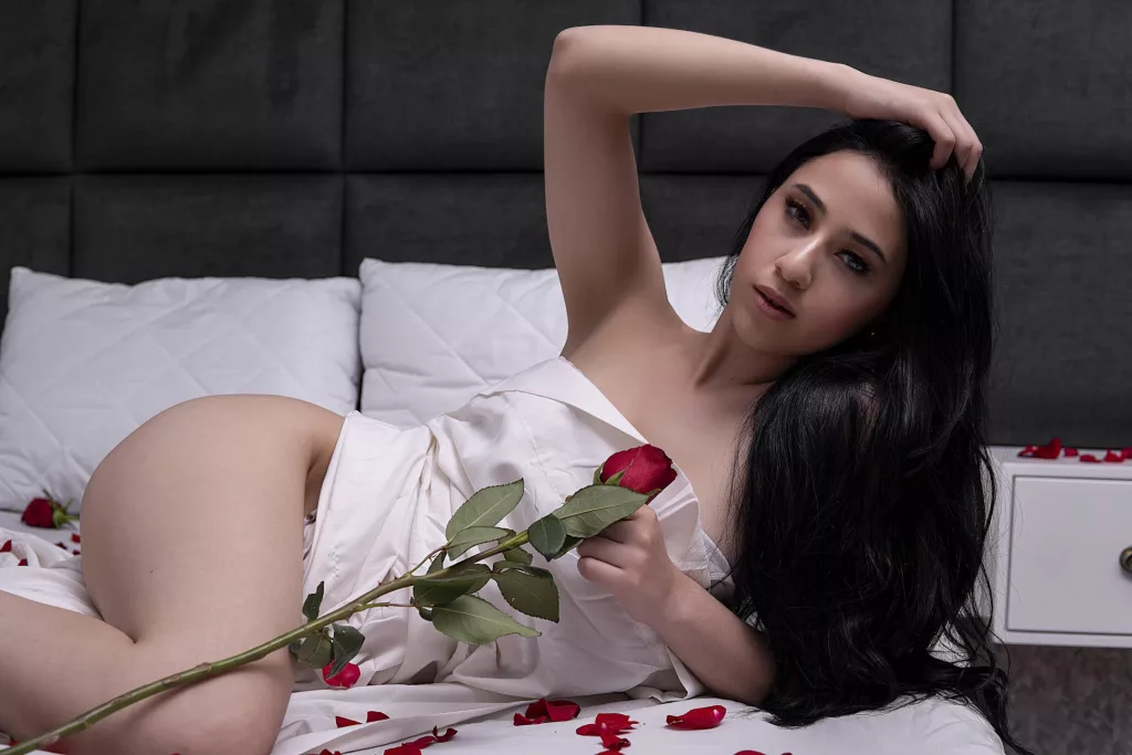 SamyCruzz lays in a all white bed with red petals and a rose at her finger tips