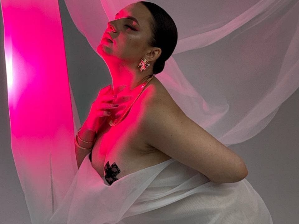 Dreia in white draped cloth, gold earrings, red-lit portrait.