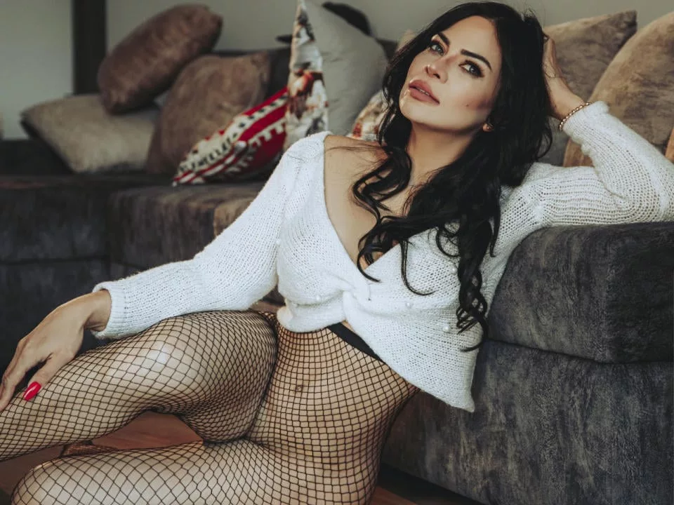 GemaHills in white sweater, fishnets, reclining on gray couch.