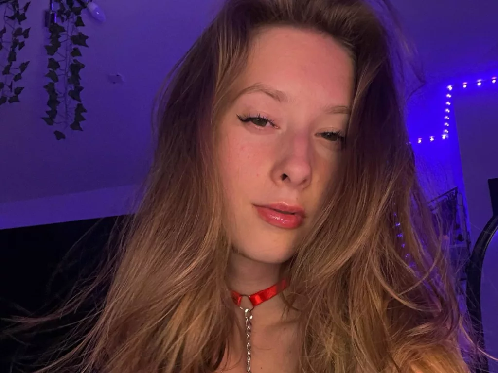 CandyFlosse, the fit girl nude, red collar, glossy lips, blue-lit background with plants.