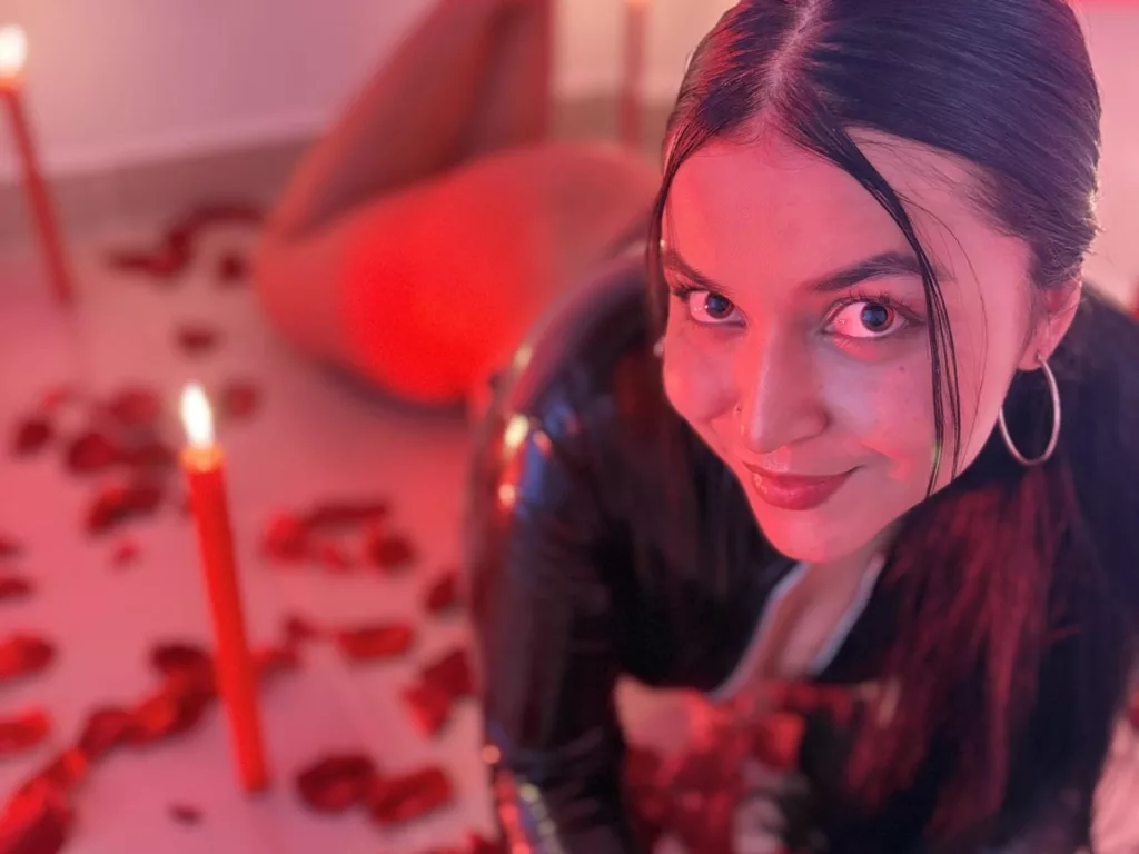 HikariChloe kneeling, mischievous grin, red candlelight and rose petals around her.