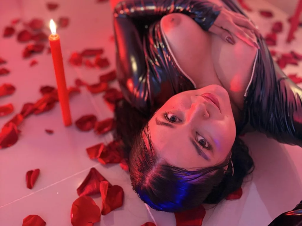 HikariChloe on red rose petals, black latex top, breasts exposed, candle nearby