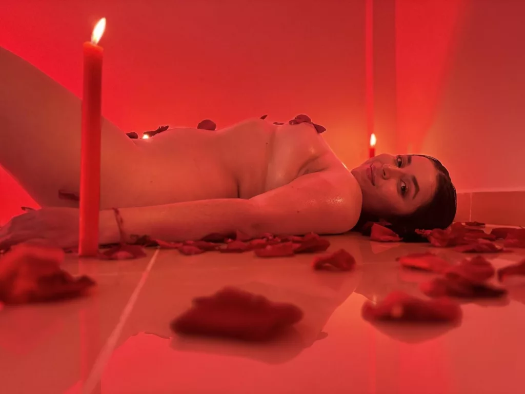 HikariChloe on red rose petals, black latex top, breasts exposed, candle nearby."