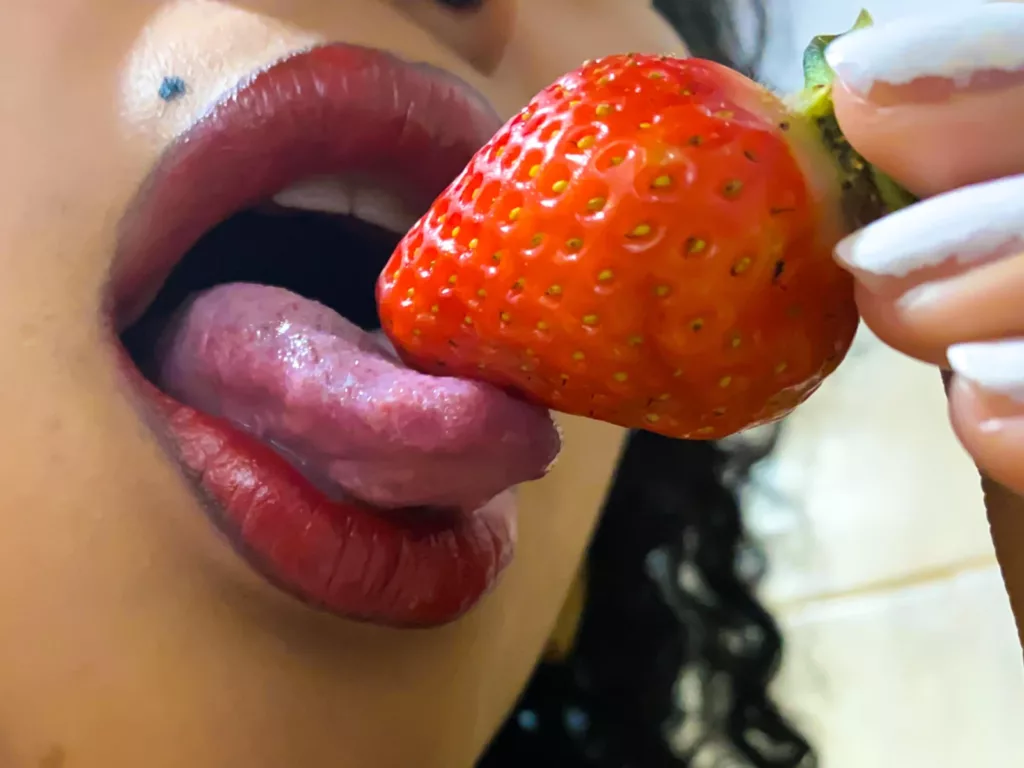SophieSonyas licks a juicy strawberry, lips glossy, tongue teasing the fruit.