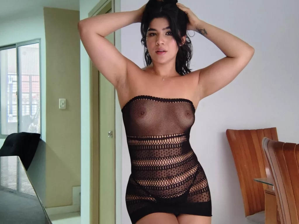 ScarlletZhen in black fishnet mini dress, arms raised, nipples exposed, modern room.
