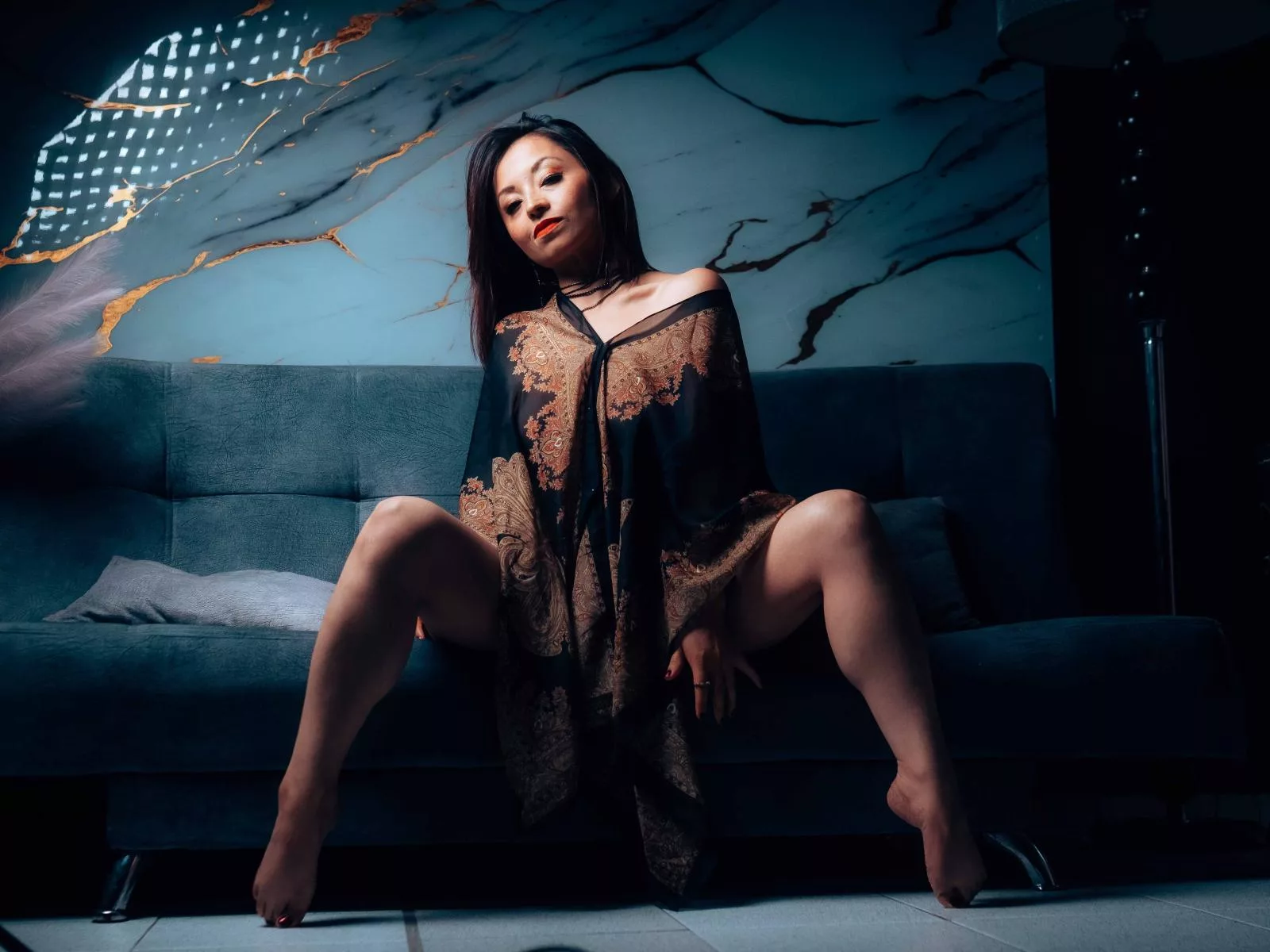 AngelinaBlanche in sheer gold dress, gray sofa, marble wall, legs spread.