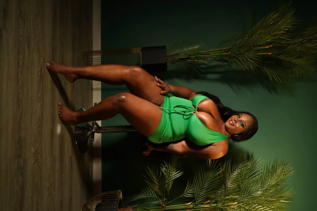 HayleyEvans in green bikini, legs apart, dark green plants, wooden floor
