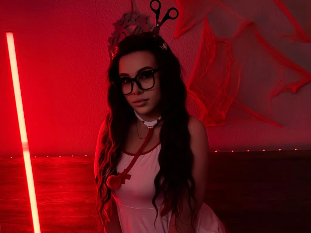 Niferttiti in a red-lit room wearing glasses and a white outfit