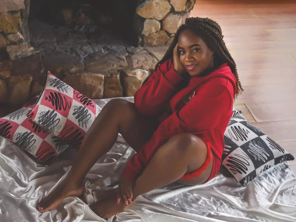 DanielaBrown, the Boss Ebony cam girl, lounges in a red hoodie and fishnet stockings on a satin bed, her playful smile inviting you into her world.