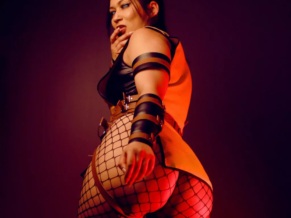 KendallBrzz mid-FDS cam model bondage show, strapped outfit with red nails under dramatic amber lighting