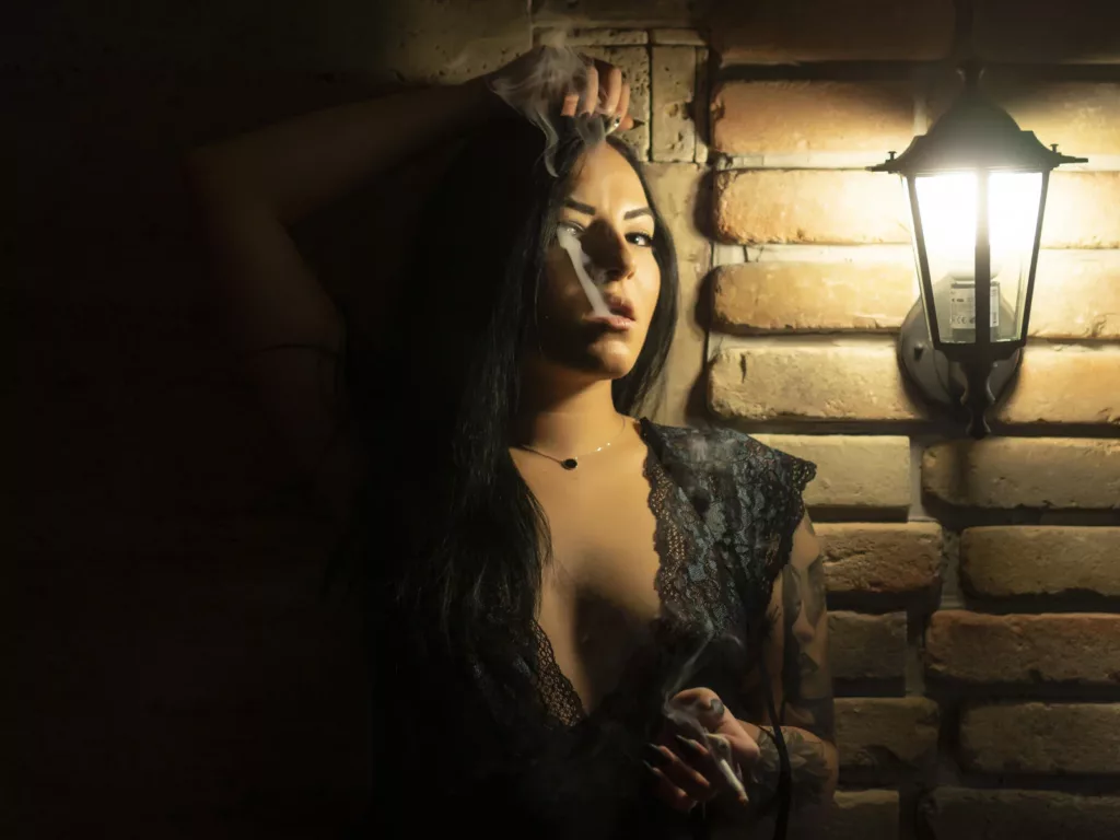 JustIzabel, the dominant BDSM Mistress, smoking seductively against a brick wall in lace lingerie