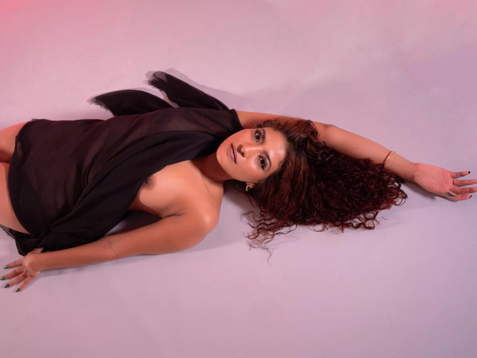 MiaBianchi, redhead cam model with long curly auburn hair, lying on her stomach in a sheer draped outfit against a pink backdrop, gazing invitingly at the lens.