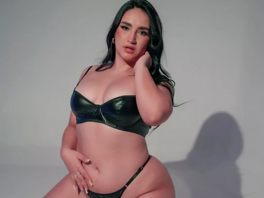MaraGrief, big boobs Latina webcam model, seated in a shiny black lingerie set, long dark hair falling over her shoulders, hand resting on her chin in a studio pose.