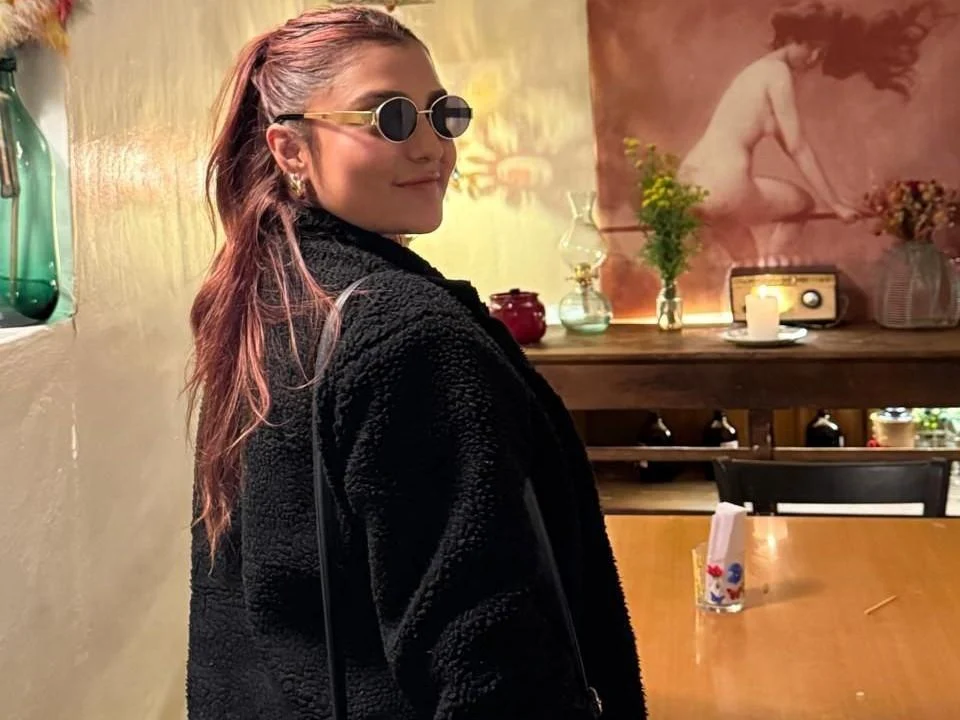 BiancaRey, curvy Colombian MILF webcam model, in round sunglasses and black coat posing in a cozy café